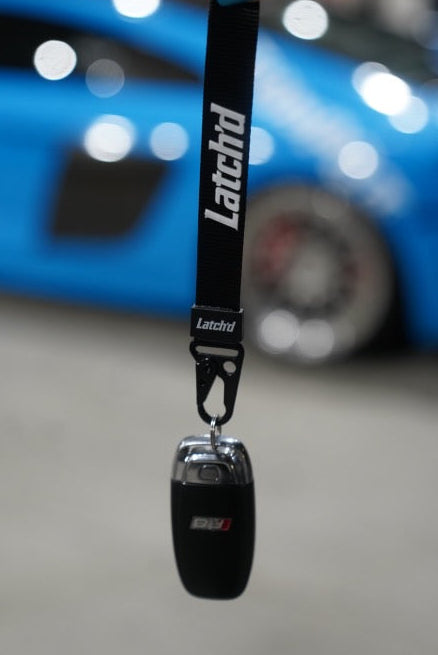 Black 'Latchtd' lanyard held by a hand with a blurred blue car in the background
